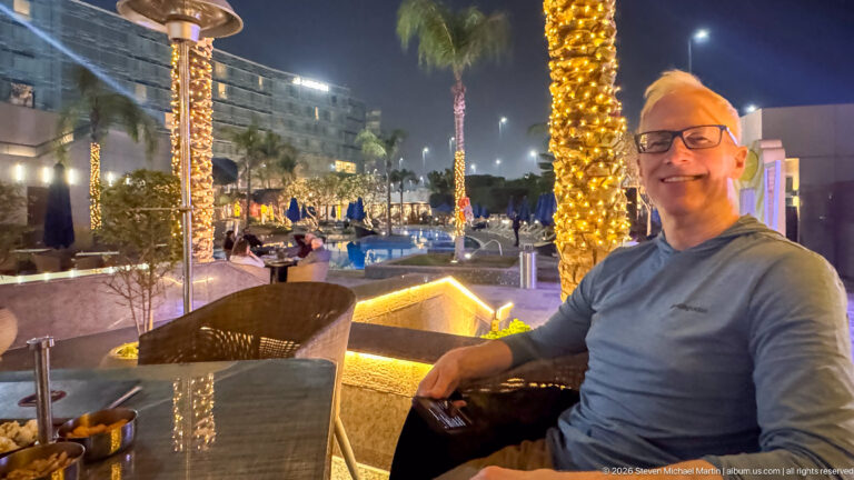 Smiling man seated at an outdoor table beside a hotel pool with illuminated palm trees at night.
