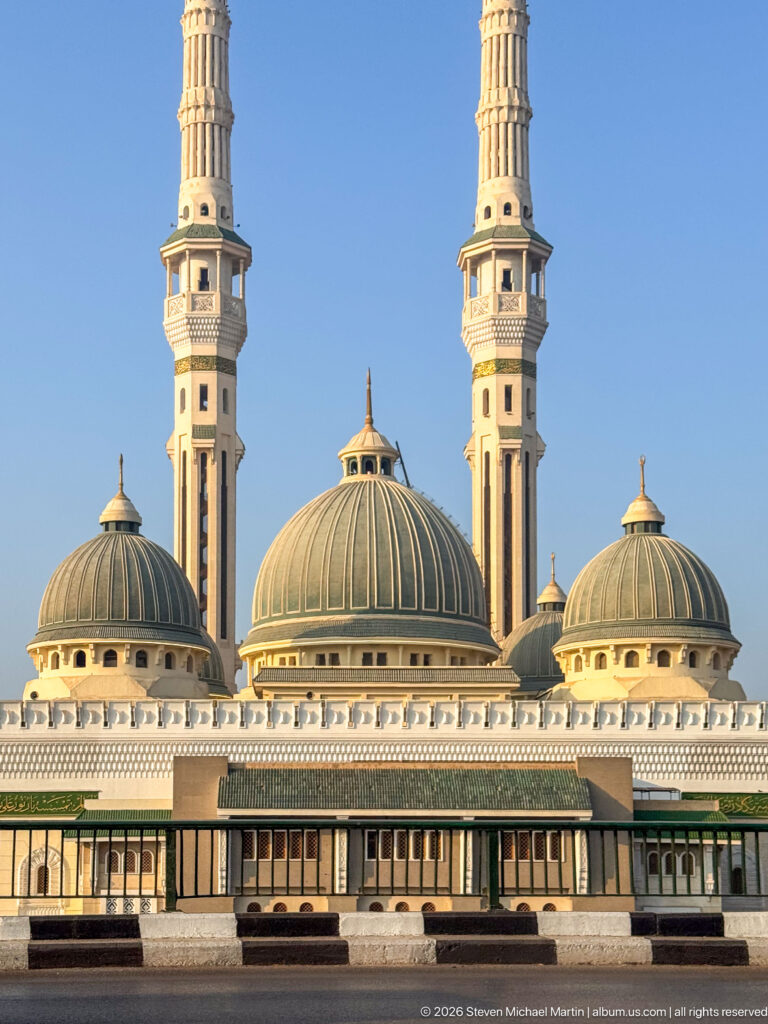 Large mosque with three ribbed domes and two tall symmetrical minarets against a clear blue sky.