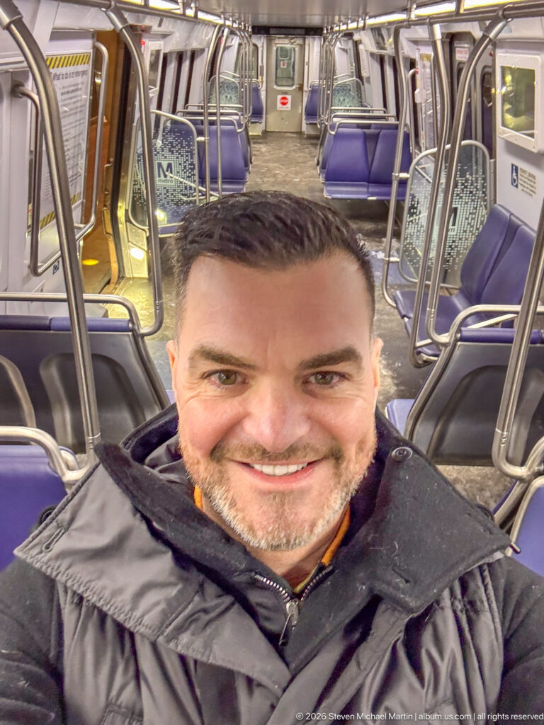 Smiling man taking a selfie inside an empty airport train car with purple seats and metal rails.