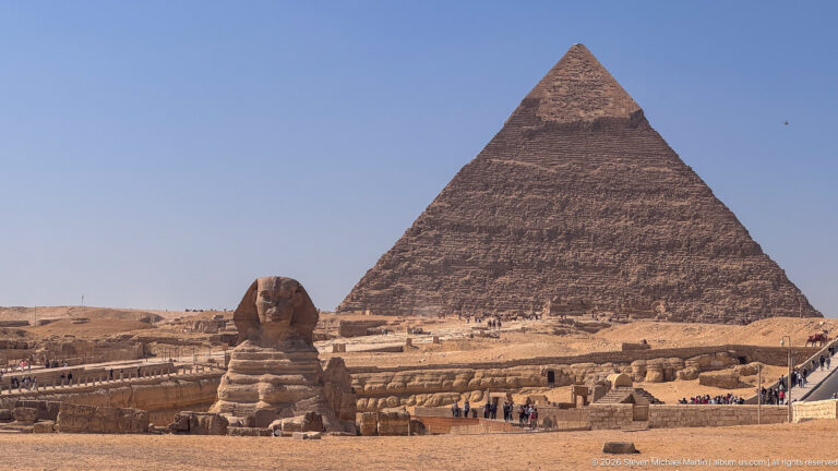 Great Sphinx centered below the Pyramid of Khafre, with visitors and excavated limestone terraces around the enclosure.