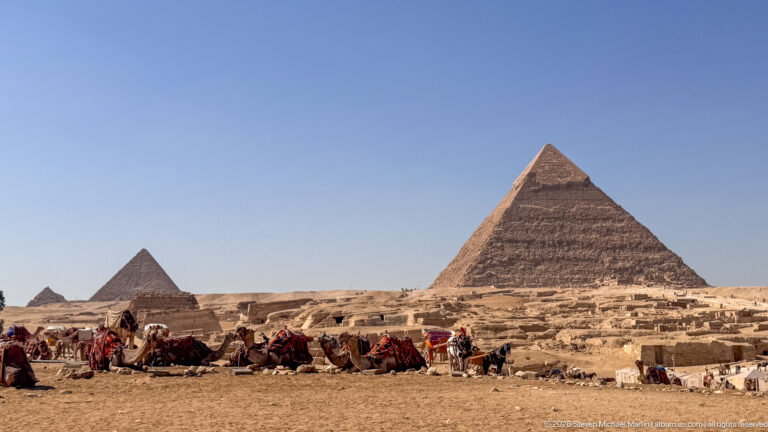 Large Pyramid of Khafre beyond saddled camels, visitors, and desert activity on the Giza Plateau.