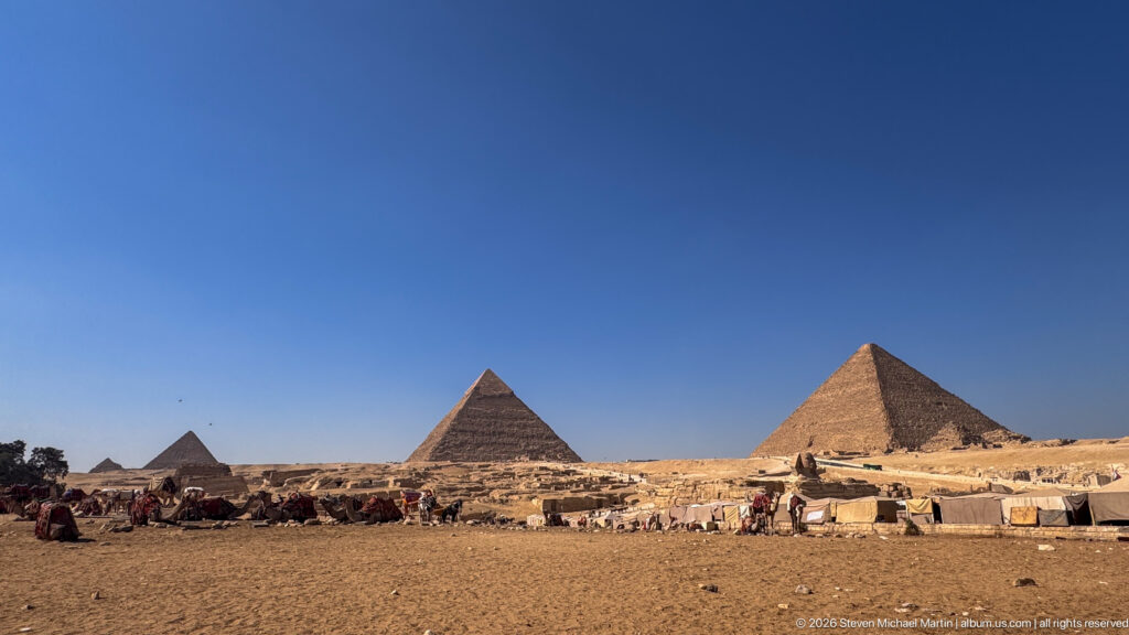 Wide Giza Plateau panorama with several pyramids, camels, visitors, tents, and open desert under blue sky.