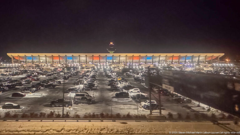 Long illuminated airport terminal at night beyond crowded parking lots and rows of bright streetlights.