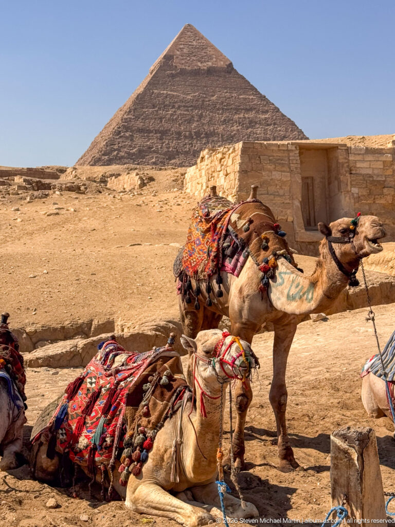 Saddled camels with colorful blankets resting beside stonework below a large pyramid on the Giza Plateau.