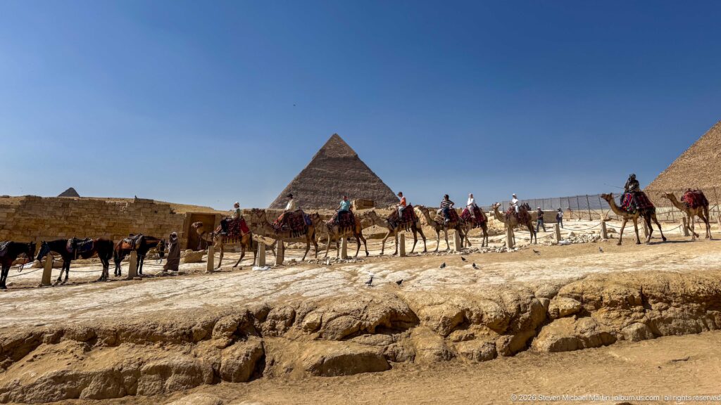 Saddled camels and riders crossing the Giza Plateau with pyramids rising in the background.