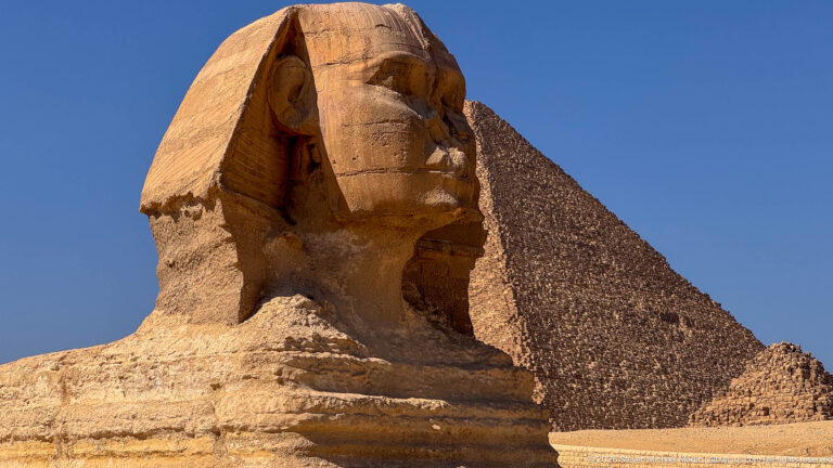 Close three-quarter view of the Great Sphinx with its weathered face and a pyramid rising behind.