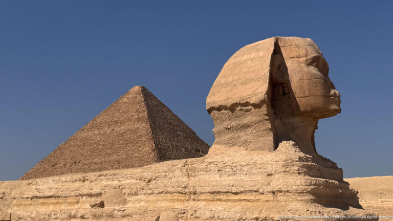 Profile view of the Great Sphinx with a large pyramid rising behind its weathered limestone body.