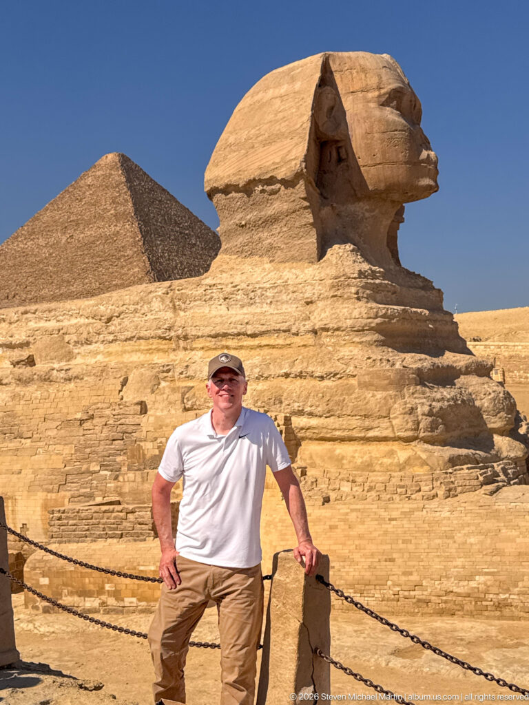 Man in a white shirt posing beside the Great Sphinx, with a pyramid visible behind the monument.