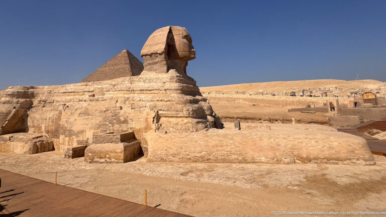 Wide side view of the Great Sphinx within its excavated enclosure, with a pyramid rising behind.