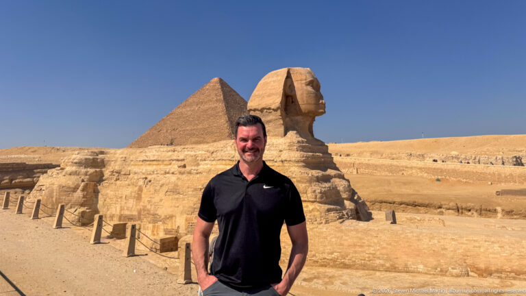 Man in a black shirt standing at an overlook with the Great Sphinx and a pyramid behind him.