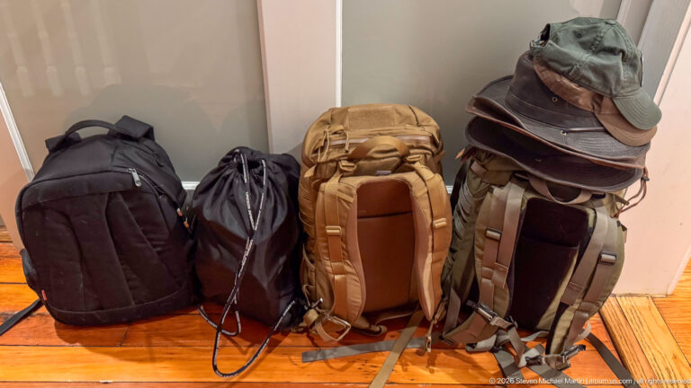 Four packed travel bags on a wooden floor, including a tan backpack and a gray pack topped with hats.