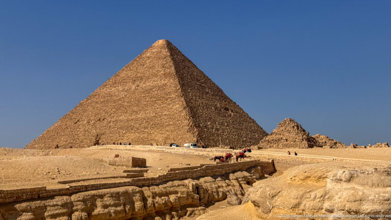 Large Giza pyramid rising above excavated stonework, visitors, and a sandy plateau under blue sky.