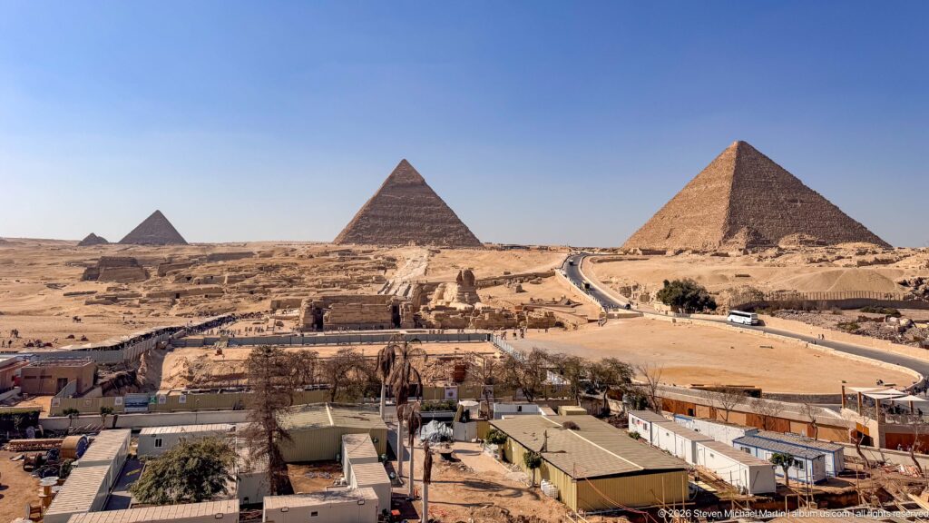 Wide elevated panorama of three major Giza pyramids across desert roads, excavations, and low buildings.
