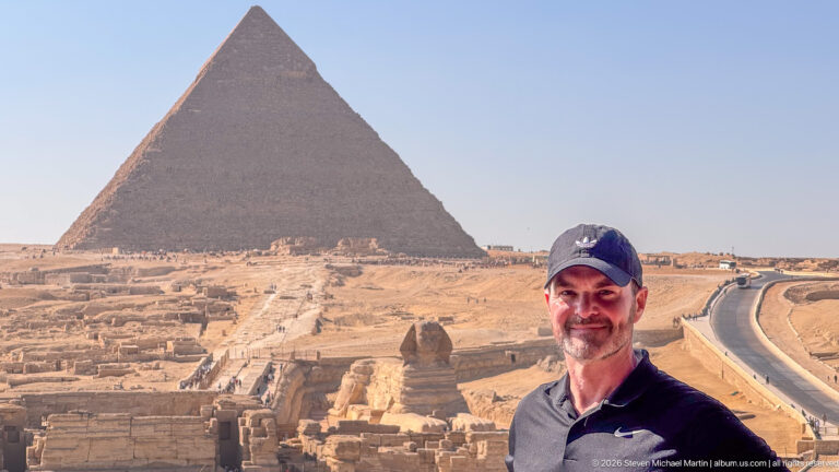 Man in dark shirt and cap at an overlook with the Great Sphinx and a large pyramid behind.