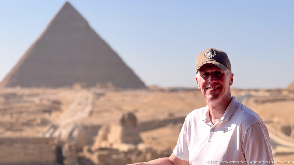 Smiling man in white shirt and cap with a large Giza pyramid blurred in the background.