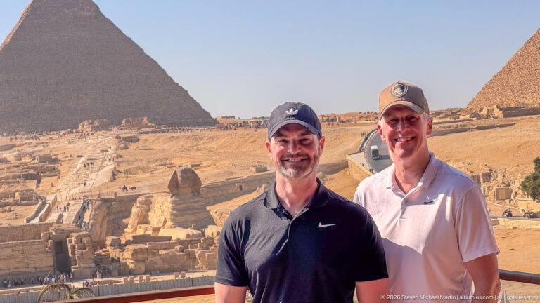 Two men posing at an overlook above the Great Sphinx with large Giza pyramids in the background.
