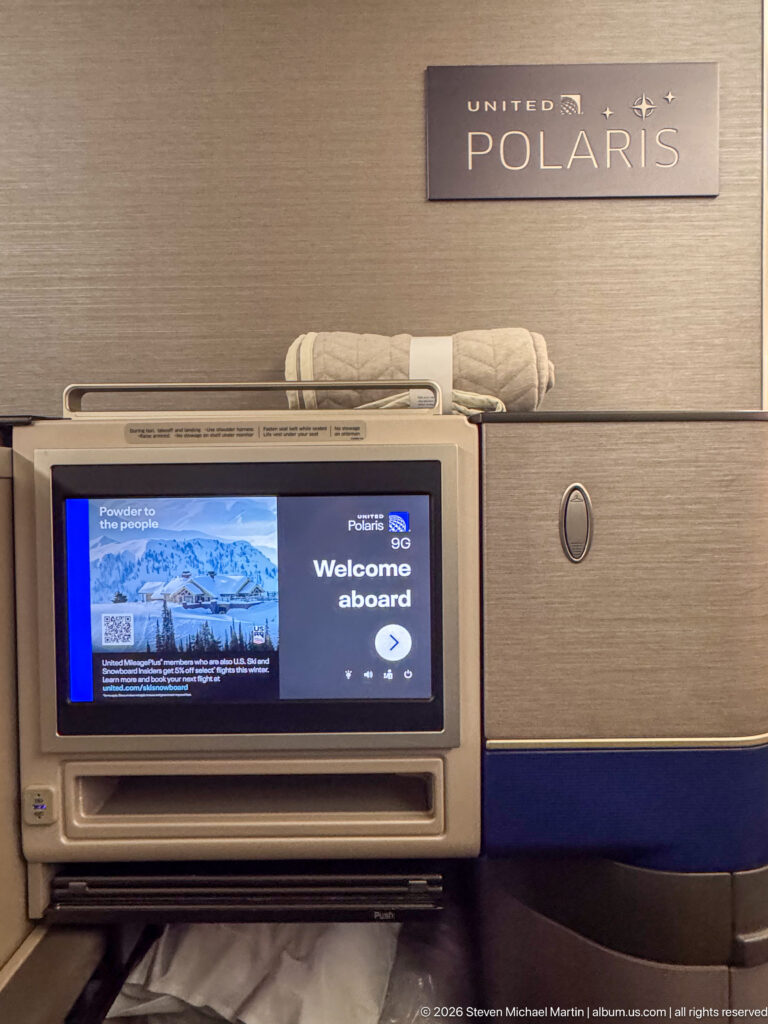 United Polaris business-class suite with welcome screen, bedding, storage compartment, and branded wall panel.