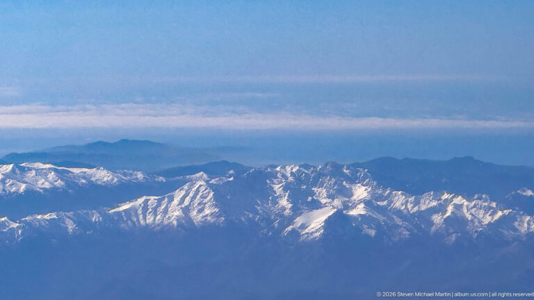 Aerial view of rugged snow-covered mountain ridges beneath blue haze and a layered sky.