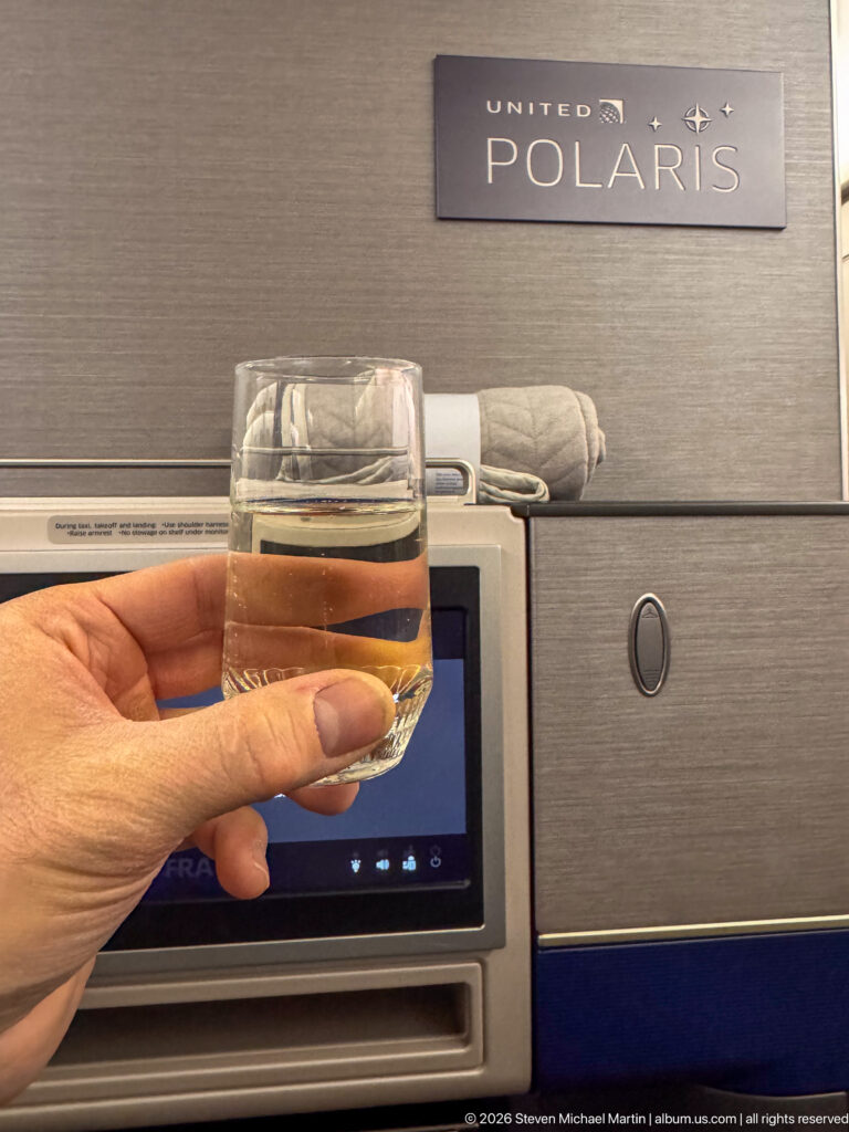 Hand holding a small glass of pale sparkling beverage in front of a United Polaris business-class seat.