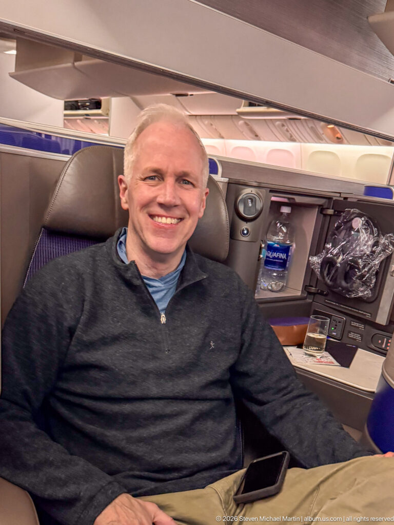 Smiling man seated in a United Polaris business-class suite with side console and bottled water.