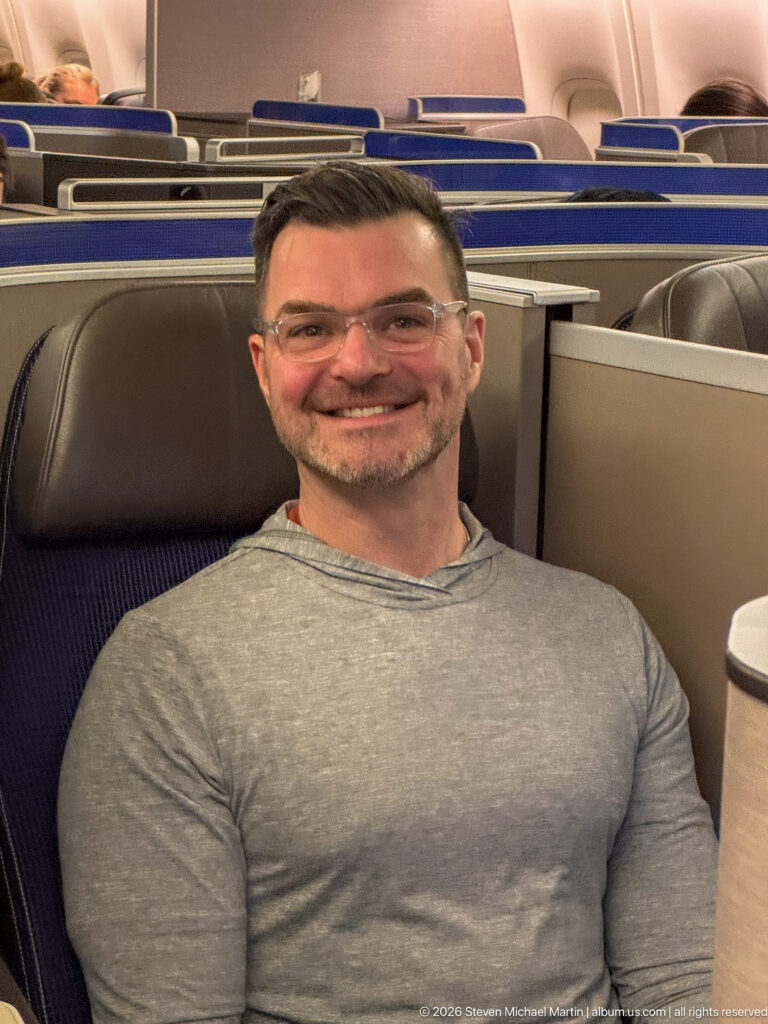 Smiling man in a gray shirt seated in a business-class pod with privacy partitions behind him.
