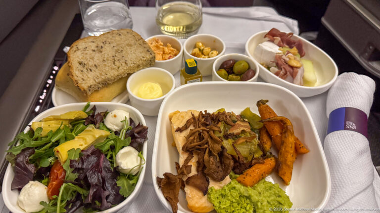 Business-class meal tray with entrée, roasted vegetables, salad, bread, olives, side dishes, and white wine.