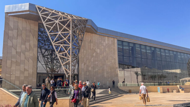 Visitors on steps outside a glazed museum entrance beneath a projecting diagonal steel lattice and pale stone façade.