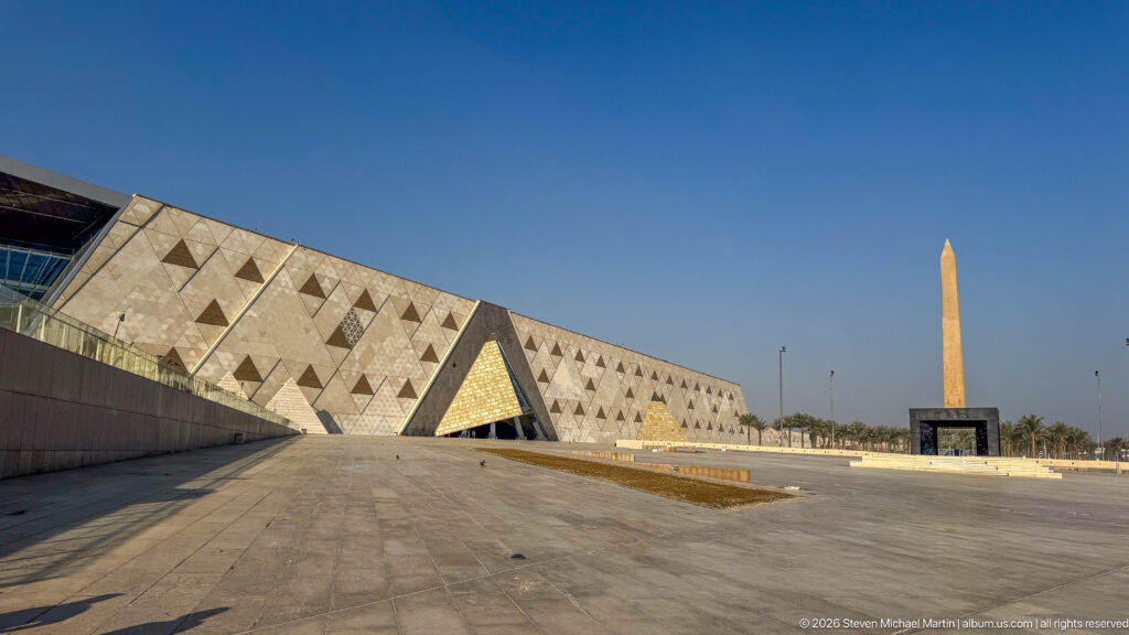 Wide museum forecourt with the long geometric façade at left and a tall ancient Egyptian obelisk standing at right.
