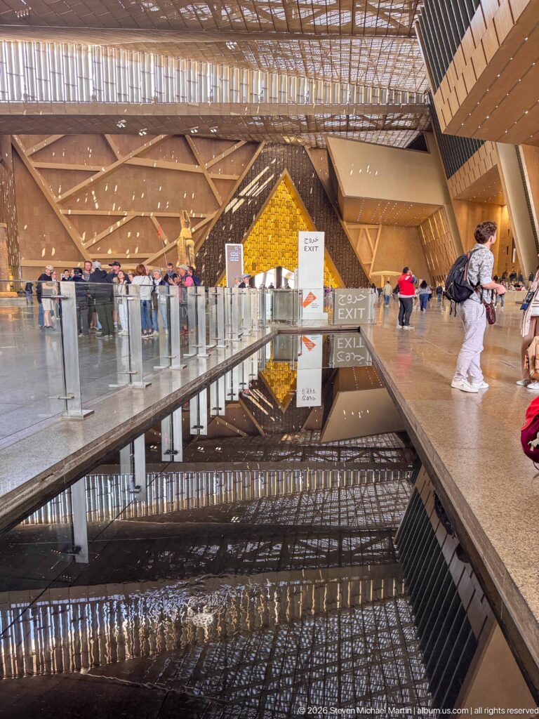 Low axial view along a reflecting pool toward a glowing triangular wall, with roof structure mirrored in the water.