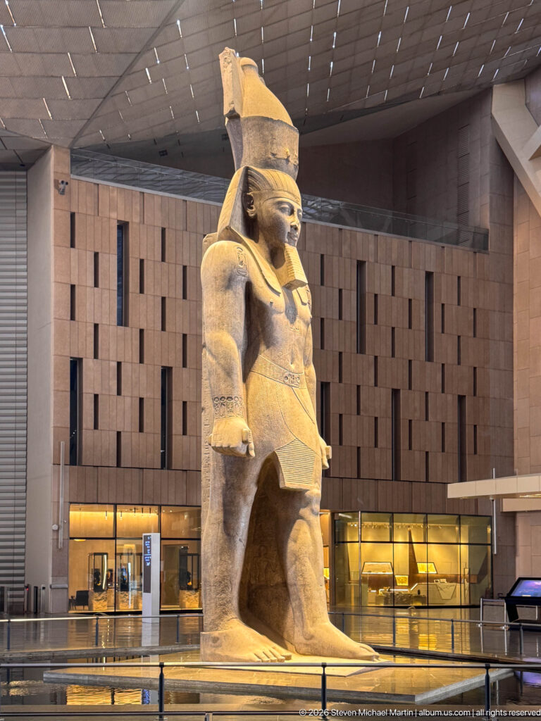 Three-quarter view of the colossal statue of Ramesses II, showing crown, forward left leg and clenched hands.
