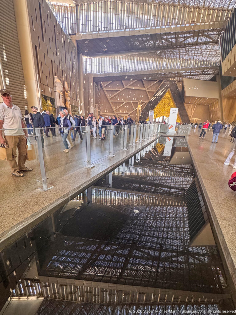 Long dark reflecting pool mirrors the patterned atrium roof, distant triangular wall, glass barriers, and visitors.