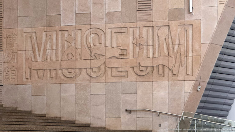 Large carved or incised MUSEUM lettering with hieroglyphic forms across a warm stone wall beside an escalator.