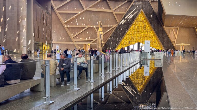 Visitors seated beside the atrium reflecting pool, with a distant statue and glowing triangular wall reflected in the water.
