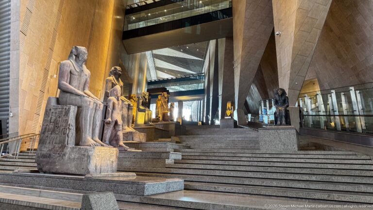 Low view up the Grand Staircase past large seated ancient Egyptian statues with hieroglyphic inscriptions on their thrones.