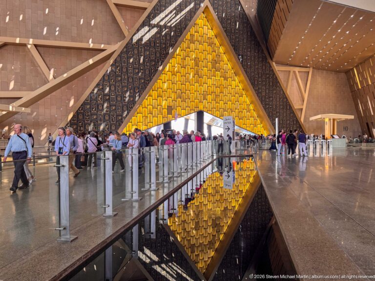 Gold-toned triangular atrium wall reflected in a long pool, with visitors and queue barriers on both sides.