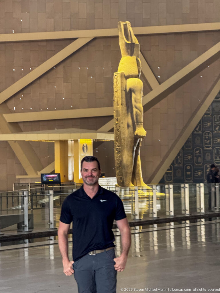 Man standing in the Grand Hall with a monumental ancient Egyptian statue behind him and angular museum architecture beyond.