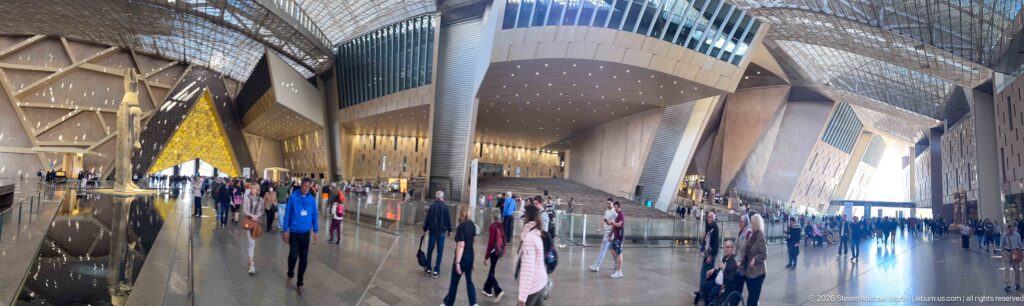 Visitors walking across the expansive Grand Egyptian Museum atrium beneath tall angular walls and a latticed roof.