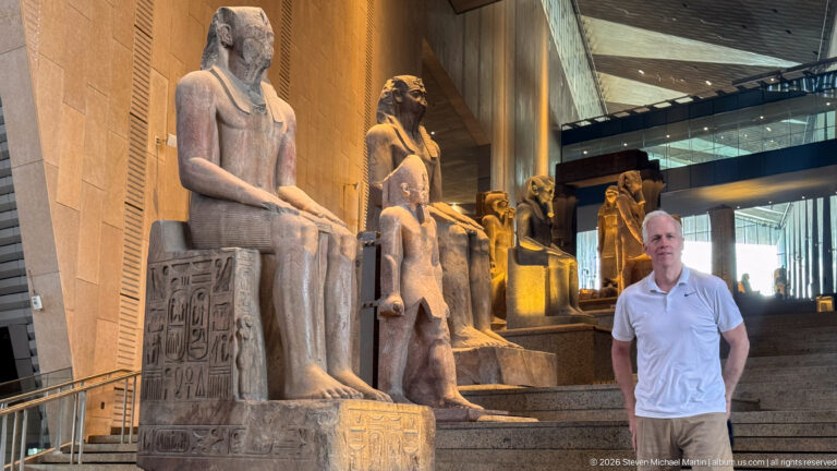 Man in a white shirt standing beside large seated royal statues on the Grand Staircase, with an inscribed throne at left.