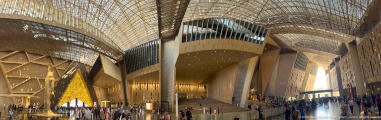 Vast Grand Egyptian Museum atrium beneath patterned roof canopy, with angular walls, visitors, and a distant monumental statue.