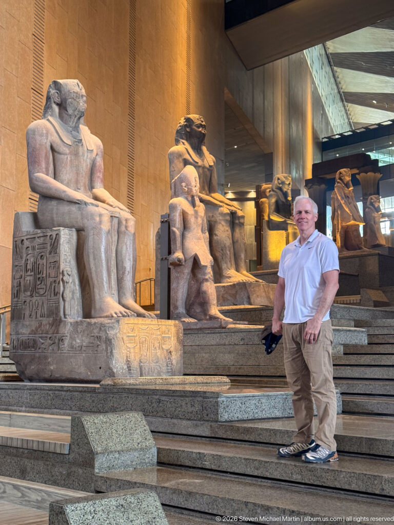 Man standing beside a monumental seated ancient Egyptian statue on the Grand Staircase, with more sculptures behind.