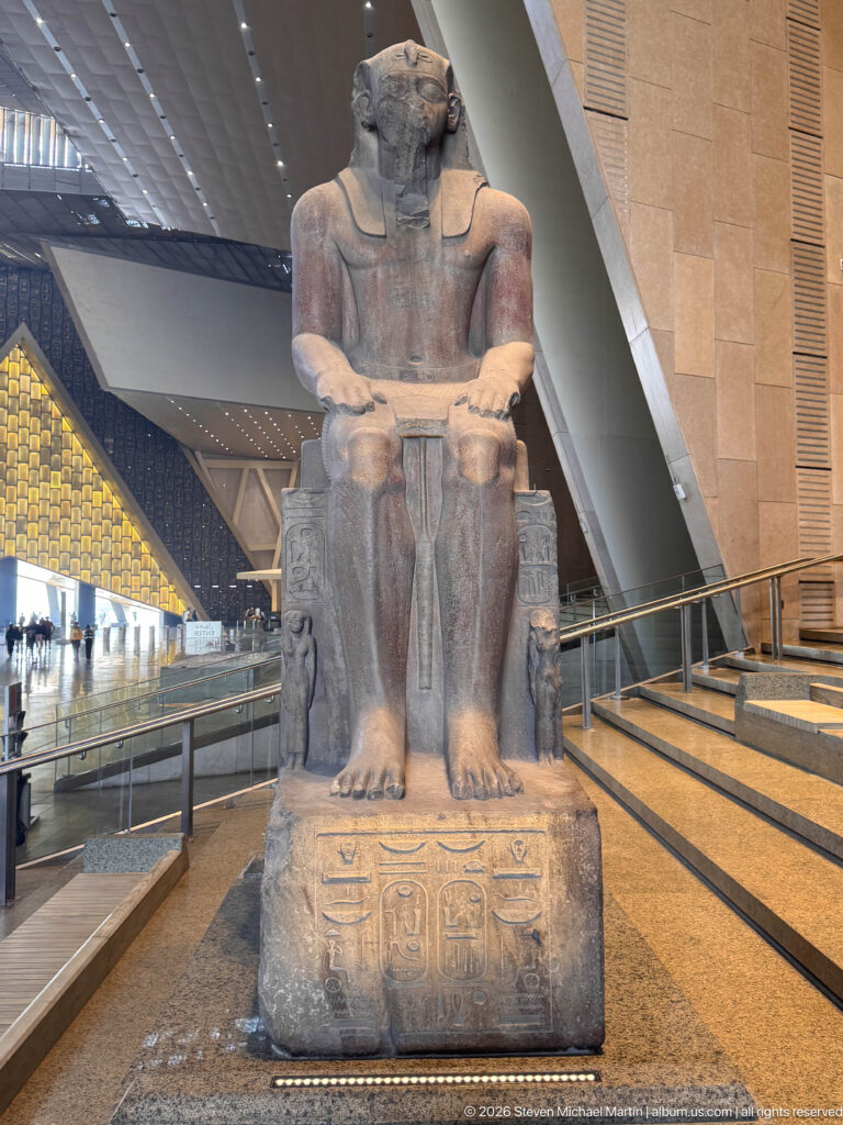 Frontal monumental seated pharaoh with hands on thighs, broad feet, and hieroglyphs carved on the throne and base.