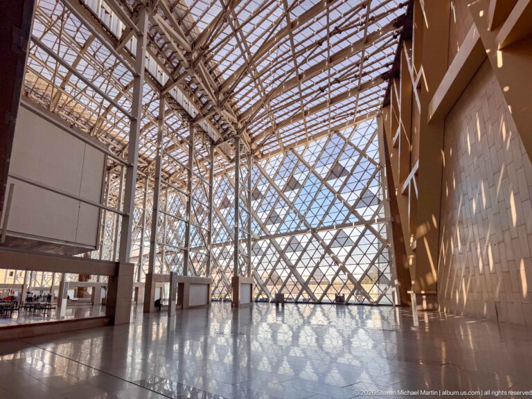 Sunlight through diagonal atrium glazing casts angular patterns onto a tall faceted wall and polished museum floor.