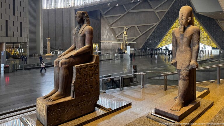 Seated and standing ancient Egyptian statues on a staircase landing overlooking the Grand Hall and bright triangular entrance.