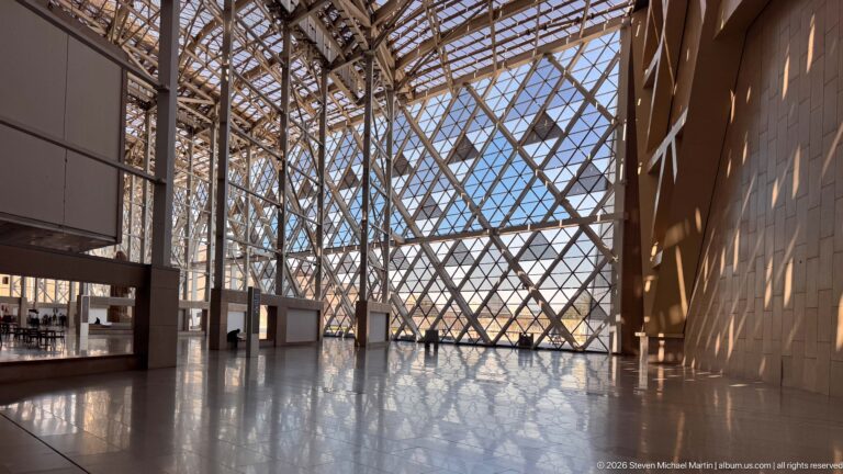 Wide atrium corner with diagonal steel lattice glazing, sunlit stone wall, and geometric reflections across the polished floor.