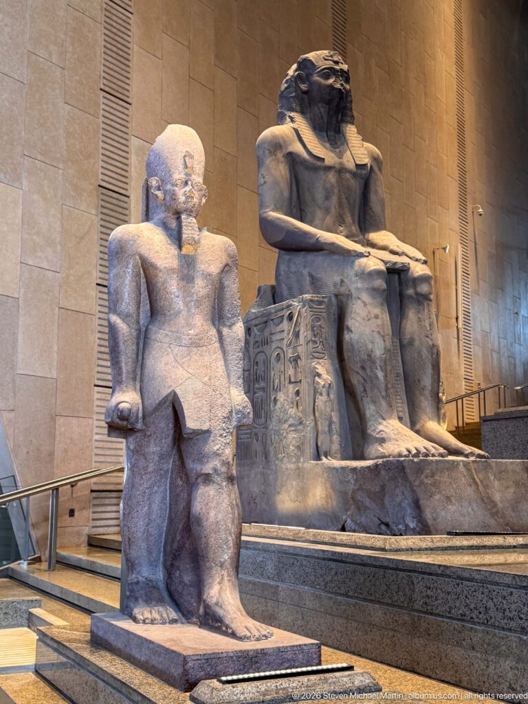 Standing ancient Egyptian male statue in front of a much larger seated royal figure on the Grand Staircase.
