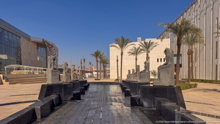 Two facing rows of seated stone figures line a narrow courtyard water channel between museum buildings and palm trees.