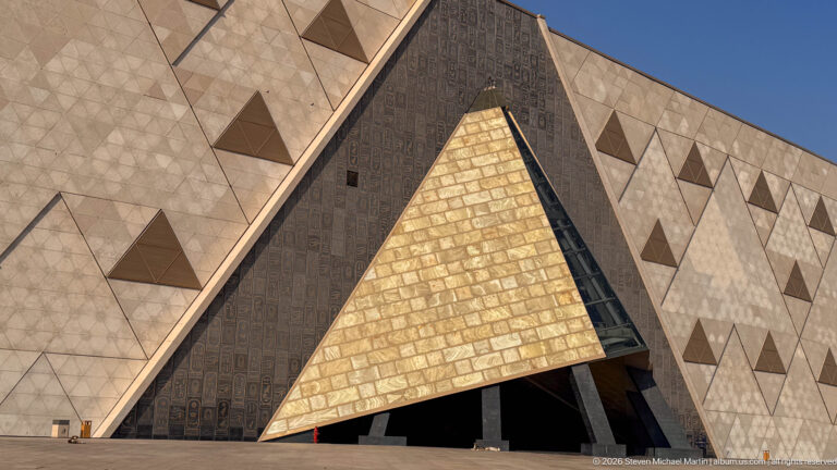 Low-angle view of the Grand Egyptian Museum façade with a large gold-toned triangular entrance framed by patterned stone walls.