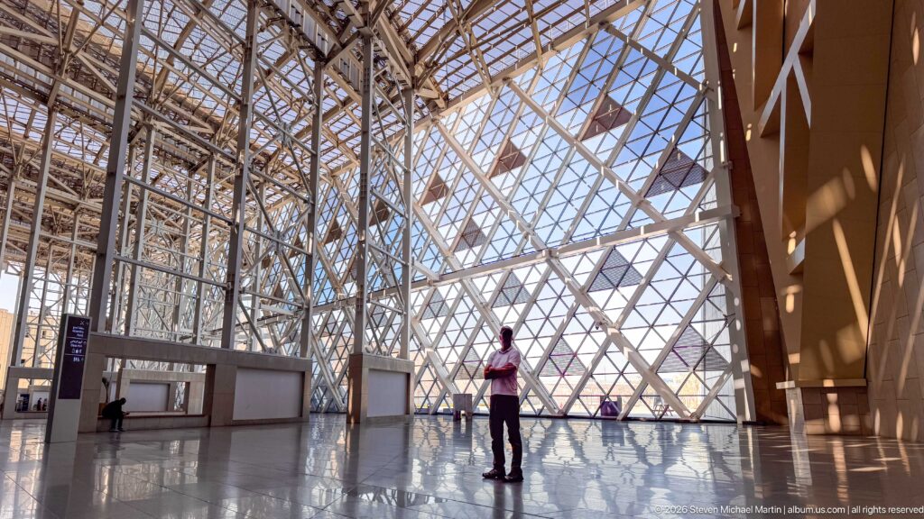 Diagonal steel lattice spans a huge glazed atrium wall and roof, reflected on the polished floor beside a visitor.