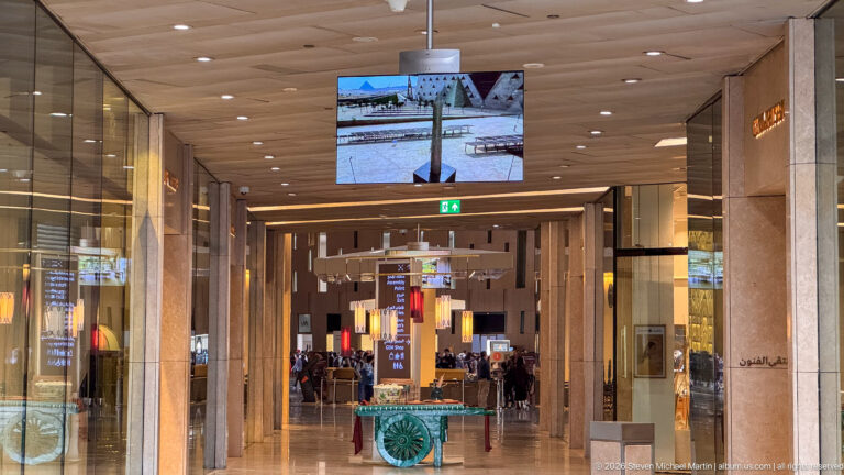 Long polished museum corridor with glass storefronts, digital screens, signs, and visitors moving through the concourse.