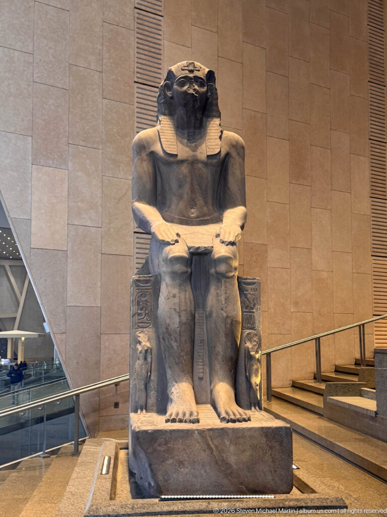 Granite seated statue of King Thutmose III wearing the nemes headcloth, hands resting on his knees above an inscribed throne.